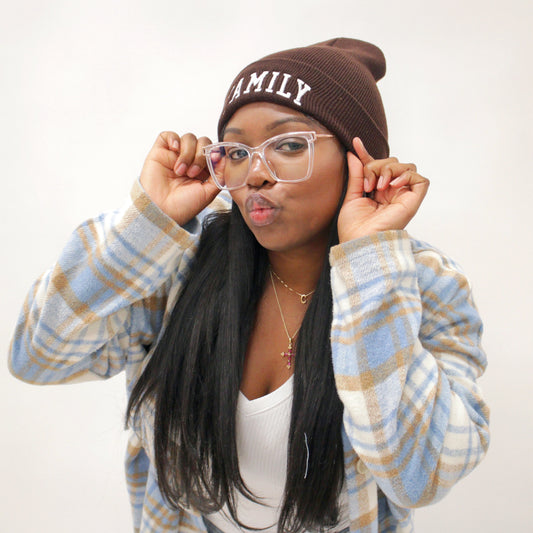 A person wearing a brown beanie with the word 'FAMILY' written on it, holding glasses with pink frames.